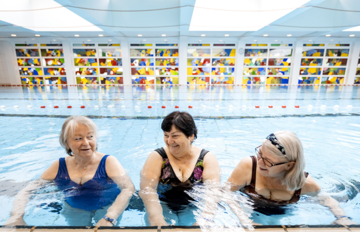Drei ältere Frauen halten sich im Therapiebecken am Rand fest, lachen und machen gemeinsam eine Reha-Übung – orthopädisch-geriatrische Rehabilitation mit sozialem Miteinander.