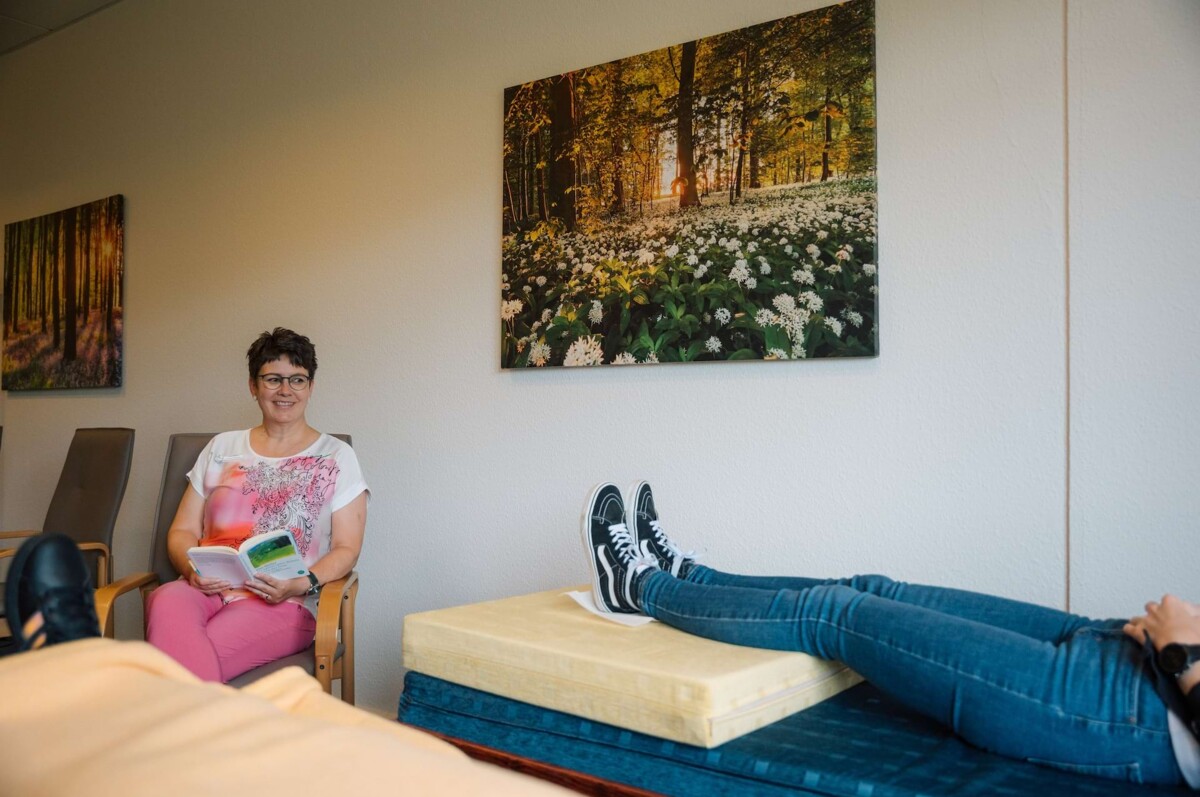 Zwei Personen im Behandlungszimmer: Eine hält eine Broschüre, die andere liegt auf der Liege – Beratung und Begleitung bei psychischen Belastungen.